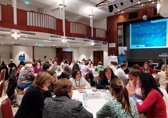 In einem Konferenzraum sitzen mehrere Personen an runden Tischen und sprechen miteinander, im Hintergrund ist der Titel der Veranstaltung auf eine Leinwand projiziert