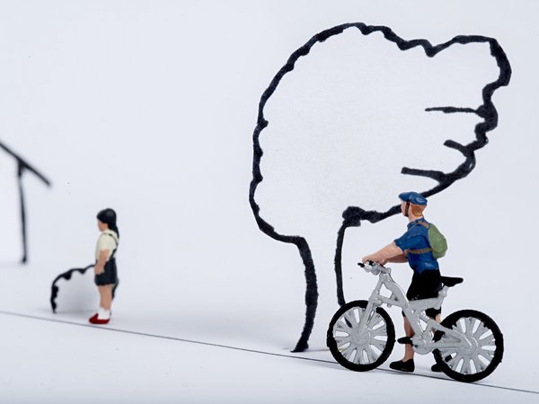Spielzeugfigur auf Fahrrad und Spielzeugfigur zu Fuß auf Straße. Gezeichneter Baum im Hintergrund. 