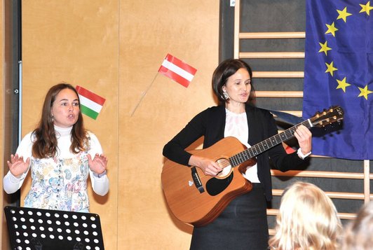Zwei Lehrerinnen stehen nebeneinander im Turnsaal. Eine hat eine Gitarre, die andere singt