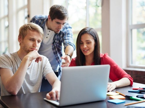 Drei Studierende schauen auf einen Laptop-Bildschirm. Zwei Sitzen, einer steht