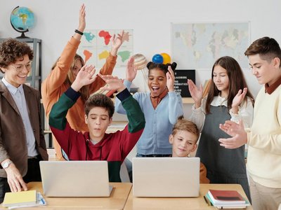 Lehrerin mit Schülerinnen und Schülern jubeln vor einem Laptop